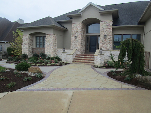 A blended red paver soldier course hugs a beautiful sandstone sidewalk, tying in varying hardscape textures and creating a striking contrast with the façade of the home.
